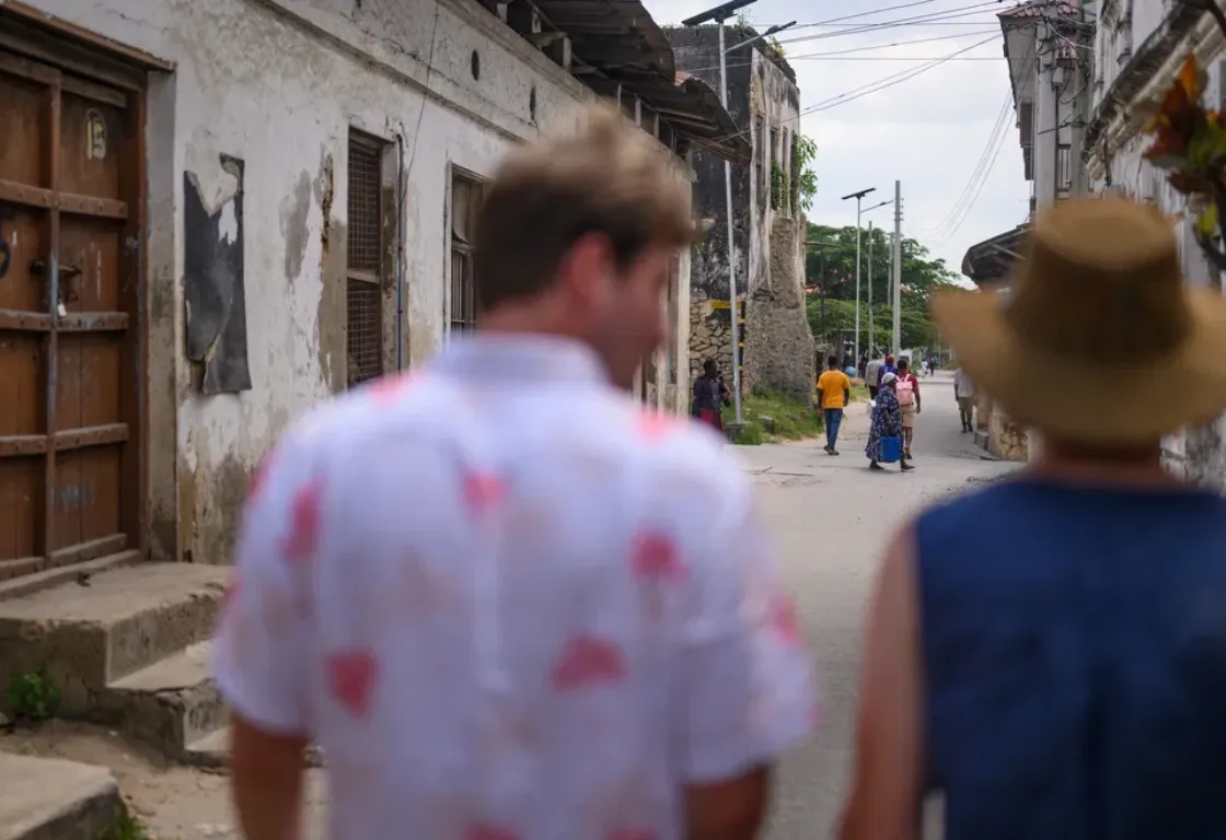 Blurred image of two people from behind walking down an old, narrow street lined with worn buildings. Others walk ahead in the distance.