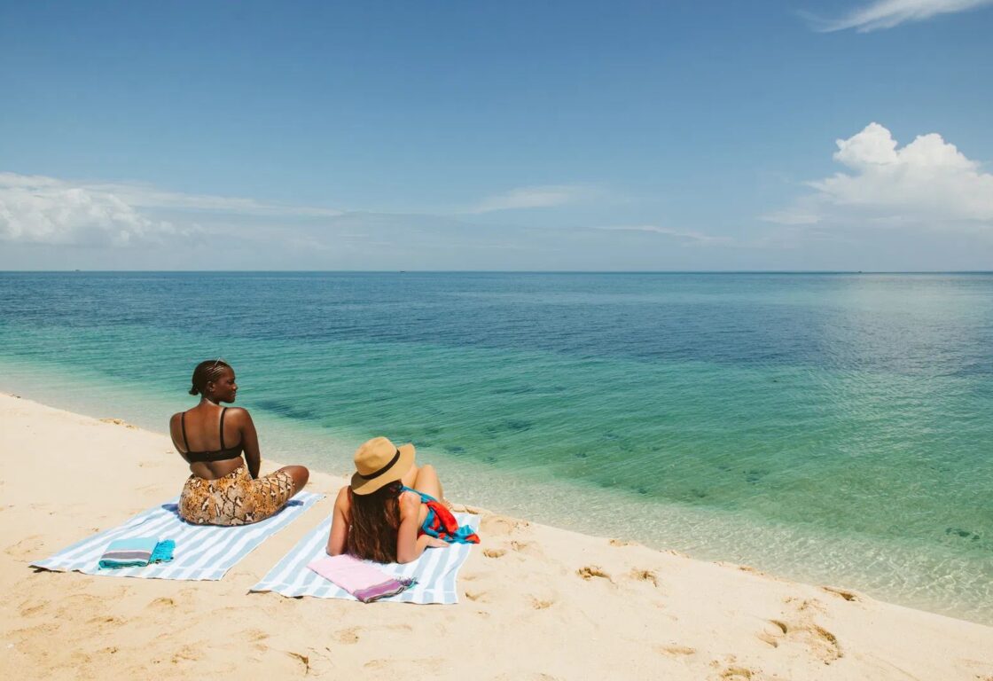 Two women relax on striped beach towels on a sandy shore, overlooking a calm turquoise ocean under a clear blue sky. The scene is peaceful and sunny.