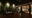 view of an enclosed wooden patio at night with a sofa against a wall covered with bamboo and leaves and on the right are glass doors and windows that look into a well lit room with sofas and foot rests