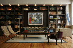 view of a wall with a book case full of books, and a variety of ornaments and in centre of the case is a large square painting of boats on water and underneath the painting is a large leather sofa, a wooden coffee table and on one side of the table are two wicker chairs and on the other is a large beige armchair and a small red armchair