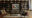 view of a wall with a book case full of books, and a variety of ornaments and in centre of the case is a large square painting of boats on water and underneath the painting is a large leather sofa, a wooden coffee table and on one side of the table are two wicker chairs and on the other is a large beige armchair and a small red armchair
