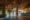 An elephant walks through a rustic lodge lobby with wooden ceilings and brick pillars. A person stands at the reception desk, lit by warm hanging lamps.