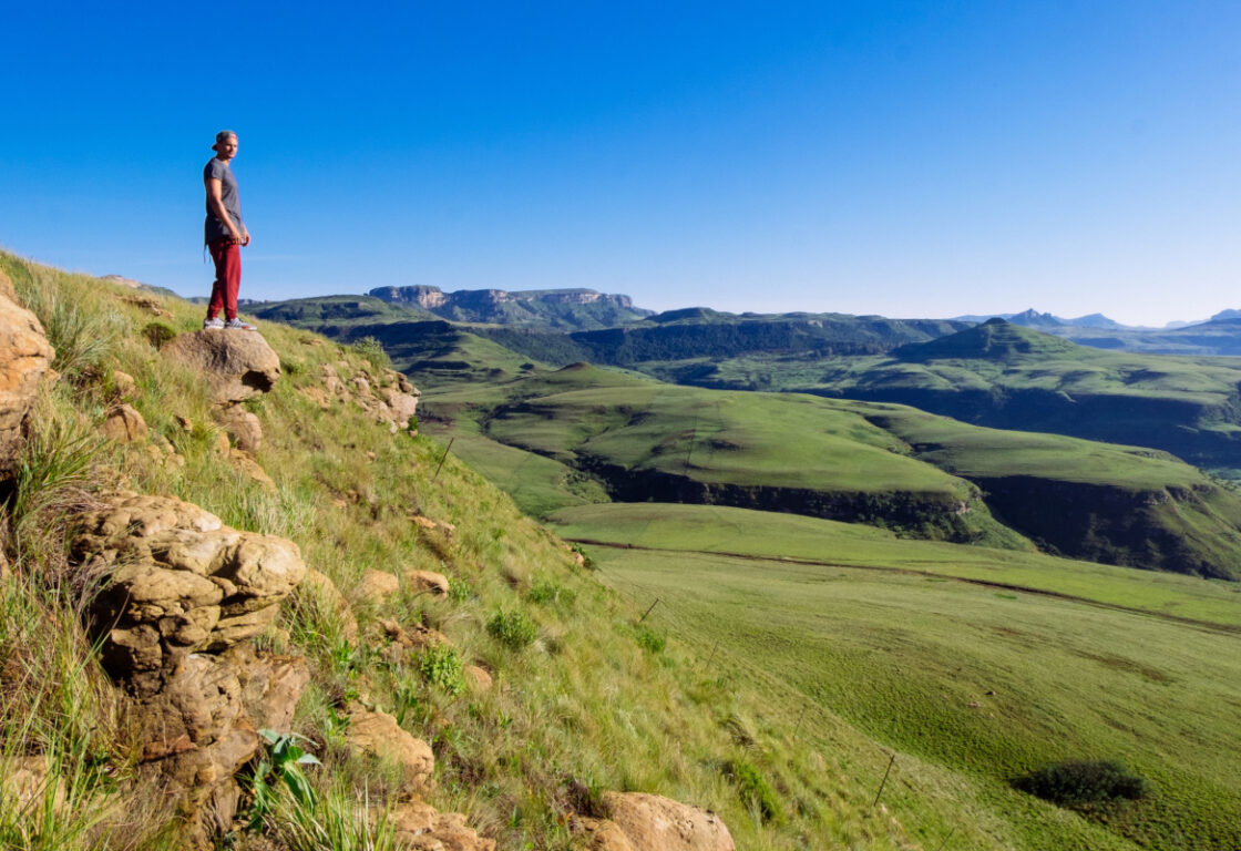 A person in a red pants and gray shirt stands on a rocky hill, overlooking a vast green valley and rolling hills under a clear blue sky.