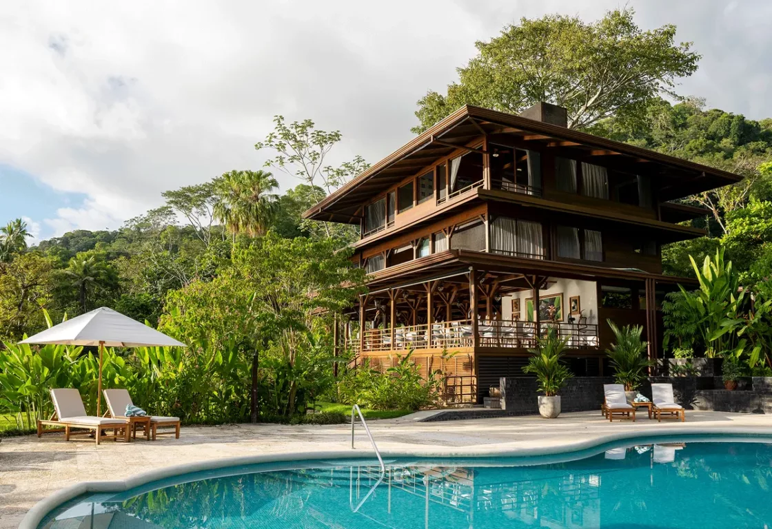 A swimming pool surrounded by a wooden building and trees, set against a clear sky with some clouds.