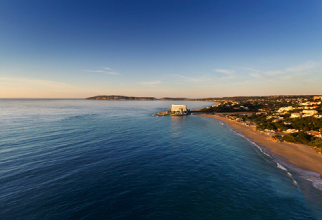 Panoramic aerial shot of the coast at sunset. Serene and calm blue water to the left, and a sandy beach fringed by green trees and houses dotted in between them, fringe the beach