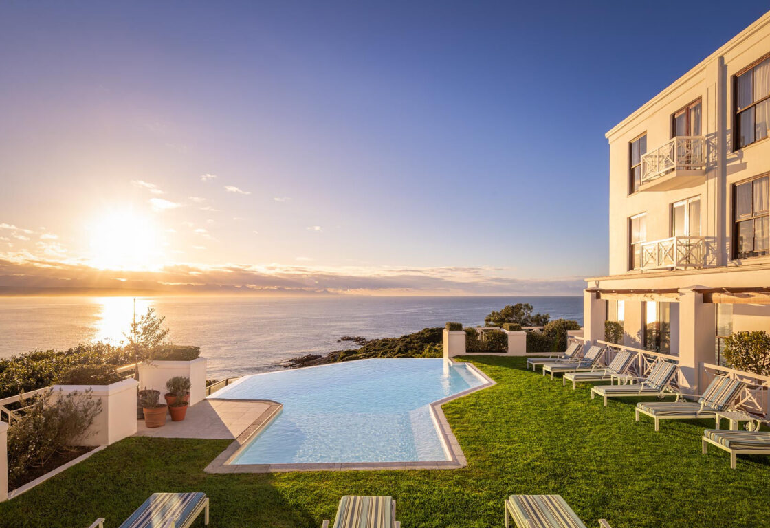 Elegant seaside resort with a stunning infinity pool, surrounded by green lawn and striped loungers, overlooking a serene ocean sunset.