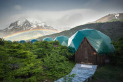 several green domes are located within Patagonian nature surrounded by mountains and trees and rainbow above it