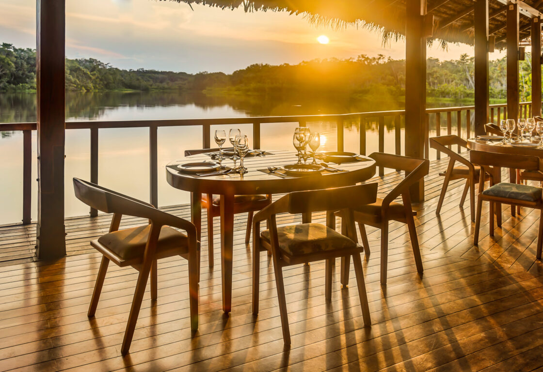 Sun is just about to set. Partial view of a wooden decked restaurant terrace, set above a river. In view is two tables with 4 chairs at each, liad out for dinner. In the background.