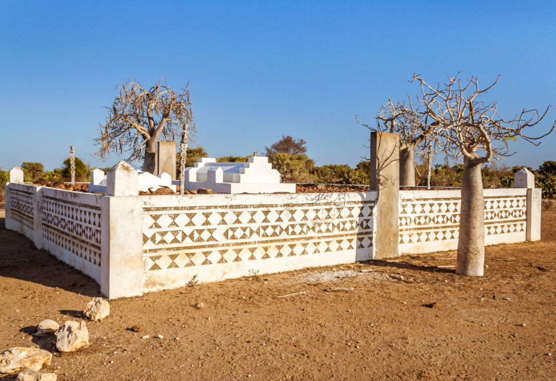 a Mahafaly burial site in madagascar shows low white walls forming a box shape and on the walls is carved patterns with brown triangles and outside the box are small baobab tees and inside are large white tombs