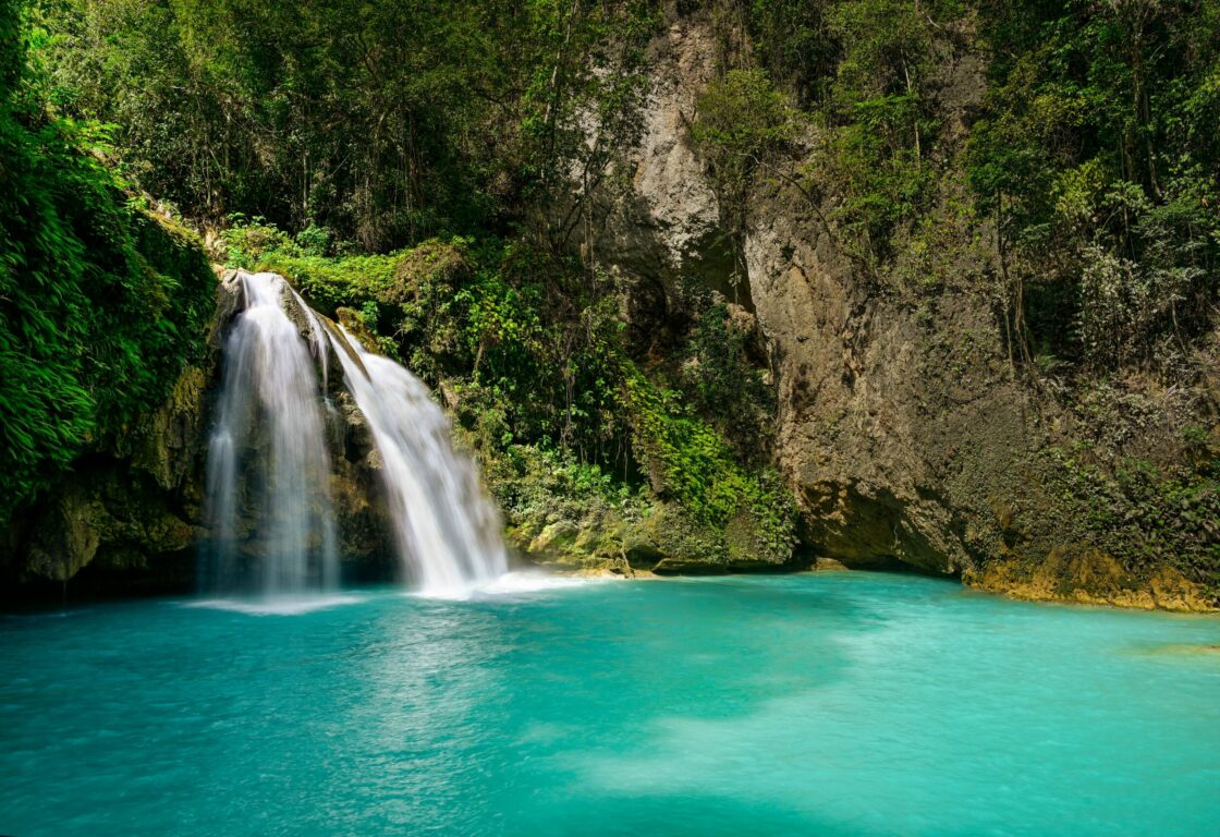 Kawasan Falls in the philippines the side of a cliff with grass and greenery growing and on the left two streams of water rush into a turquoise pool at the bottom