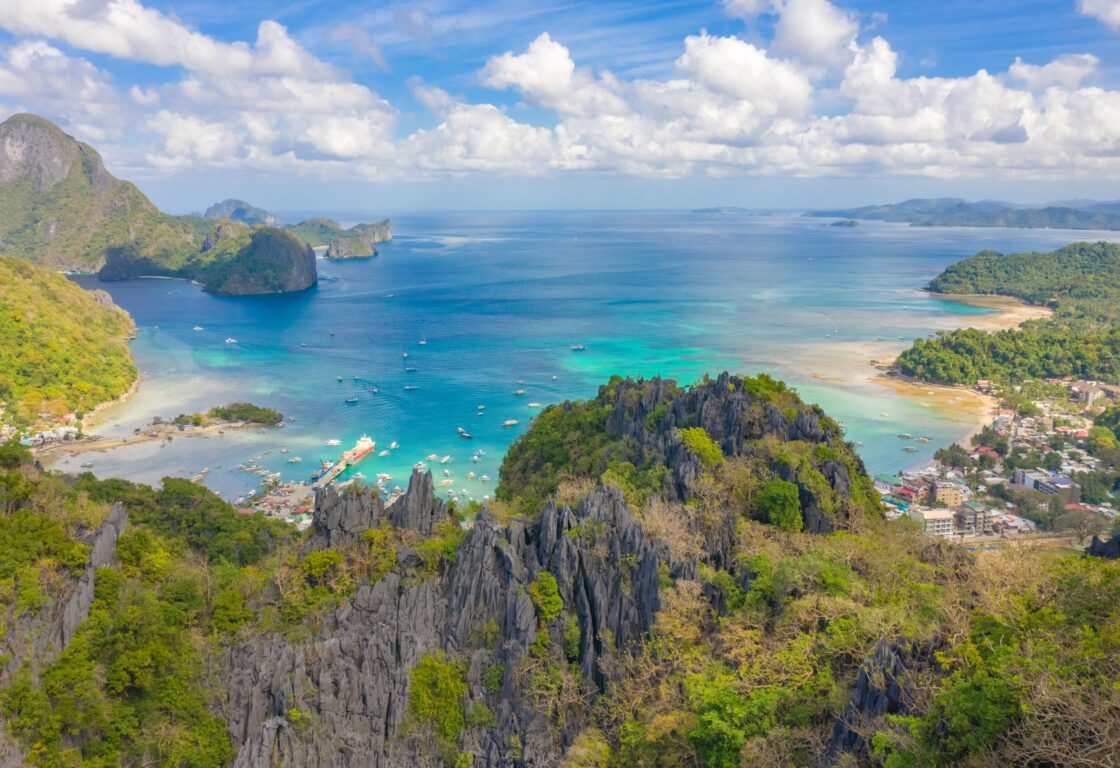 picture taken from a viewpoint in el nido in the philippines that shows jagged limestone cliffs and beyond that is sea in different shades of blue with boats and where the sea meets land there is a cluster of buildings