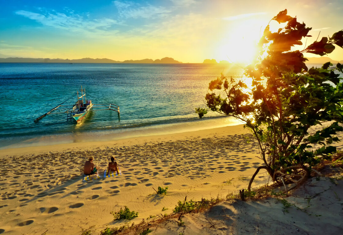 a man and woman sit on a beachwith perfect golden sand and looking at a calm sea with a traditional boat on and the sun is golden in the sky