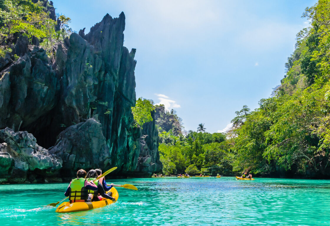 a group of 3 people sit in a yellow kayak on turquoise blue water heading towards a lagoon created by limestone cliffs and in the background there are more kayaks heading in the same direction