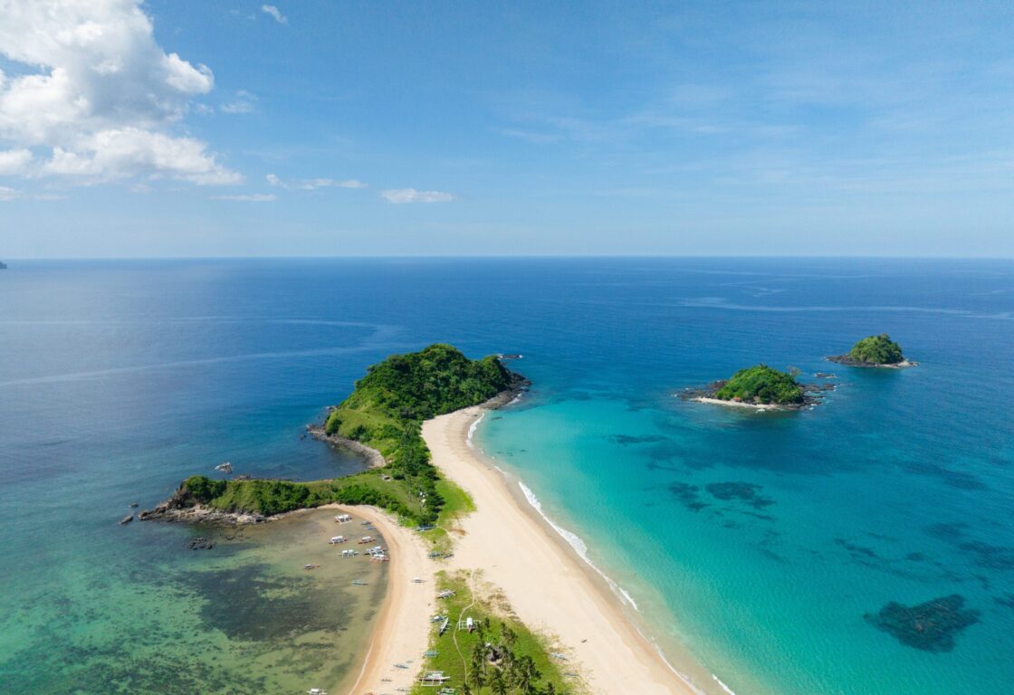 an aerial view of nacpan beach in el nido, philippines, shows a stretch of white sent with a clump of trees at the end going out into a blue sea and little boats are parked along one side of the sand