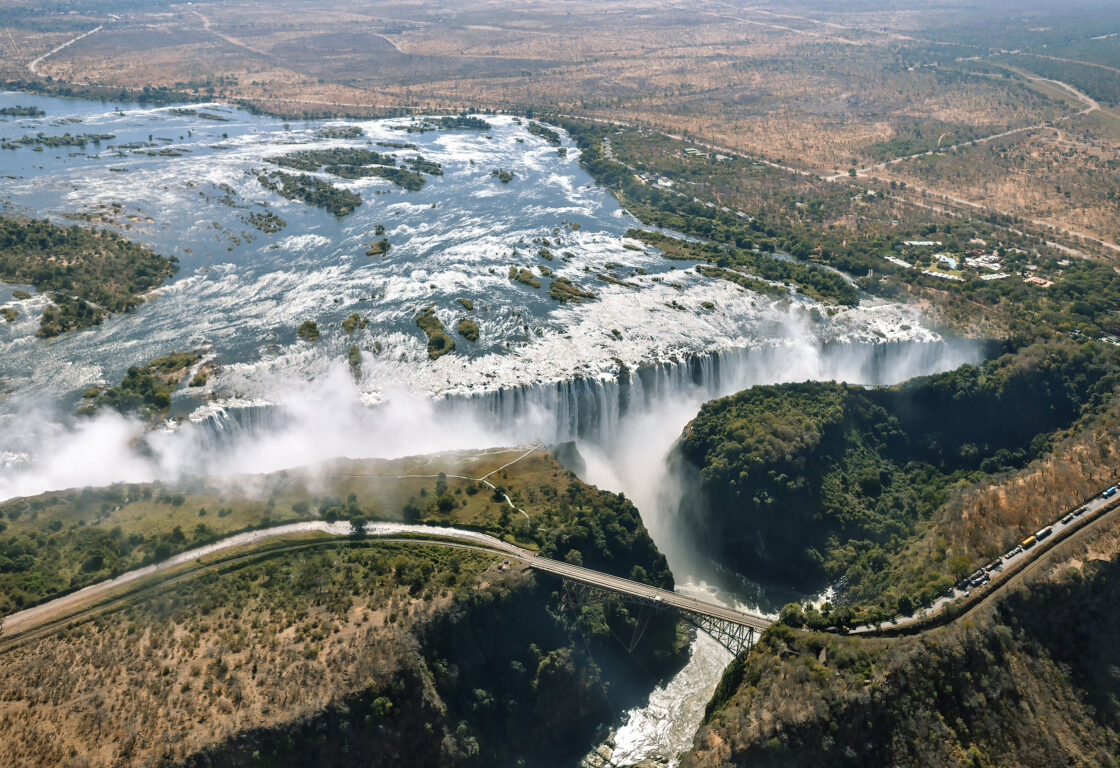 Aerial shot of the incredibly wide Victoria Falls, with the water from the Zambezi River turning white before cascading down and creating so much spray that it resembles smoke rising up from the mouth of the waterfall.