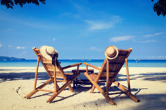 two wooden chairs on a white sanded beach face a blue sea and on each chair there is a sun hat and in the sea boats are dotted around