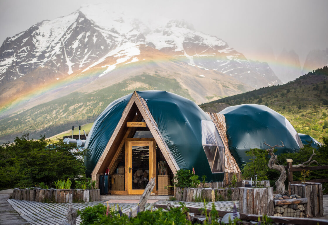several green domes are located within Patagonian nature surrounded by mountains and trees and rainbow above it