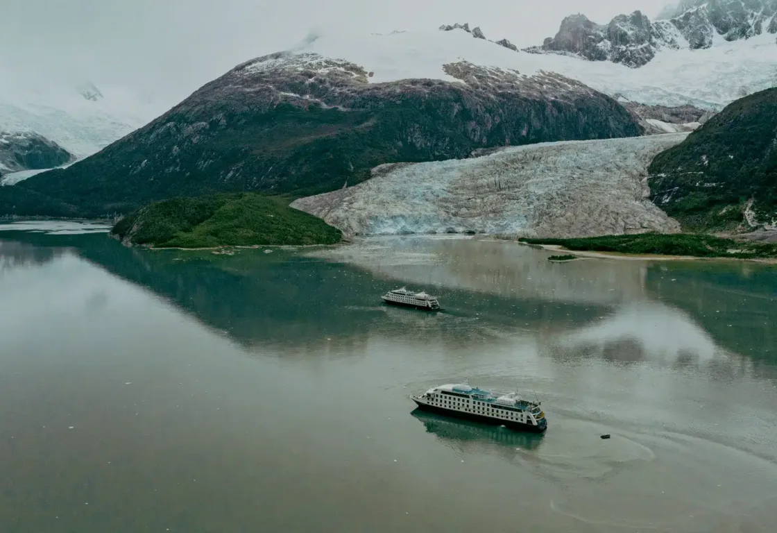 Dramatic aerial image of two csmall cruise ships sailing through a fjord while huge snowy mountains rise up around the water.