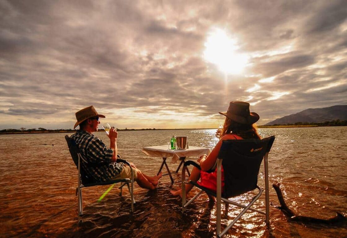 a couple siting in the shallow waters of a river bank with a table and sundowner drinks looking at the sunset