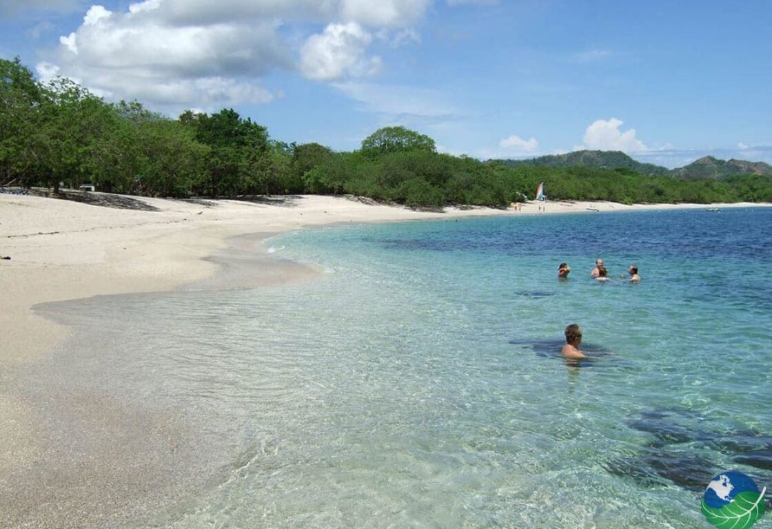 A white sandy beach stretching round into a bay with shimmering blue turquoise sea. On the borders of the beach are lush green tropical trees.
