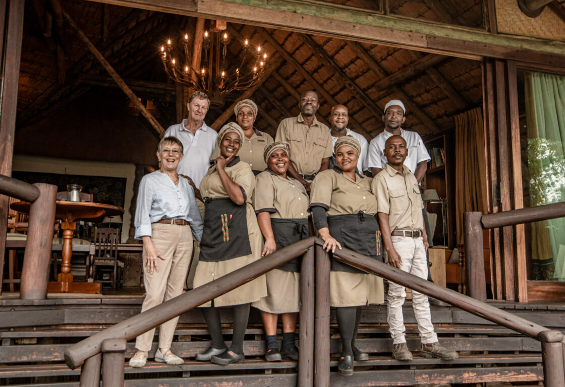 A group of lodge staff and two guests stand smiling on the wooden steps of a rustic thatched-roof building. The group includes men and women in a beige uniform.