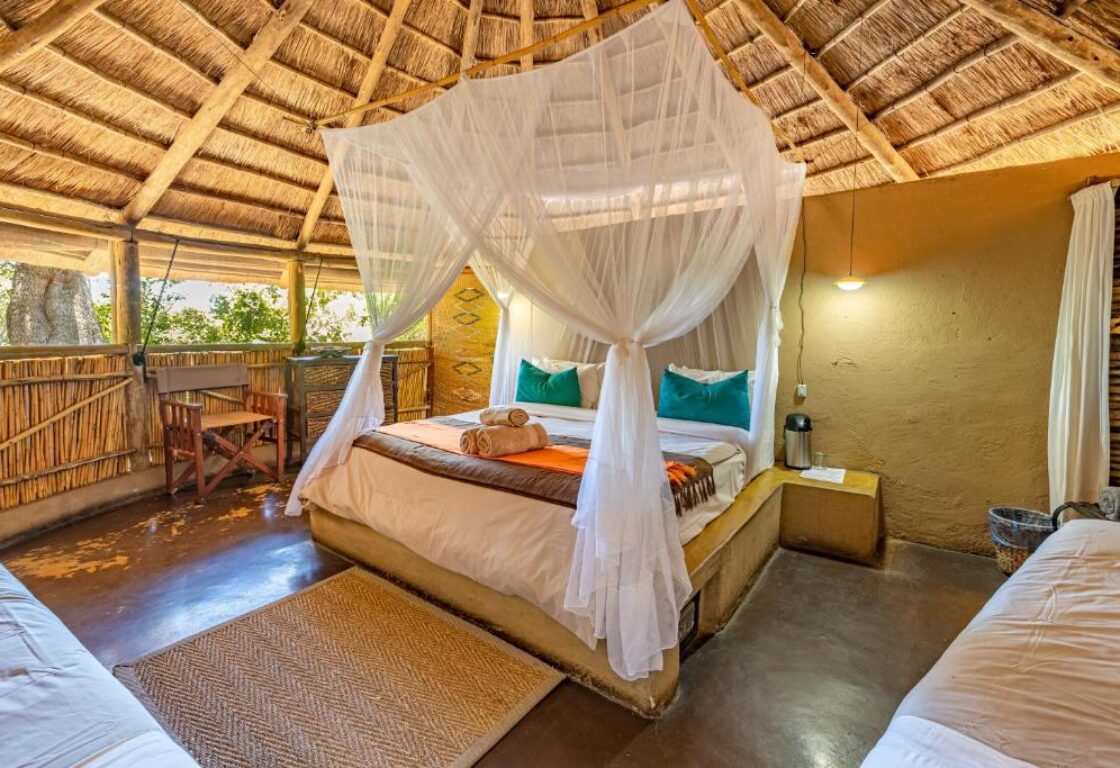Rustic bedroom with a thatched roof, featuring a four-poster bed draped with mosquito netting. Warm tones, bamboo walls, and a cozy, inviting atmosphere.