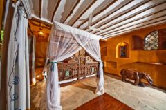 a view of a bedroom in wooden style, large bed with white sheer curtains, white small rugs, wooden hippo decoration