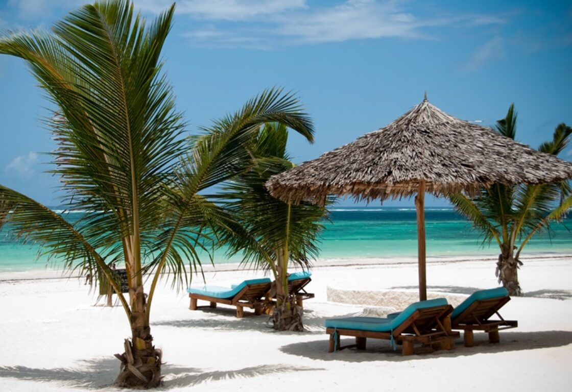 a view of a stunning white sandy beach and turquoise water with loungers featuring blue cushions and a sun brolly made of dry straw