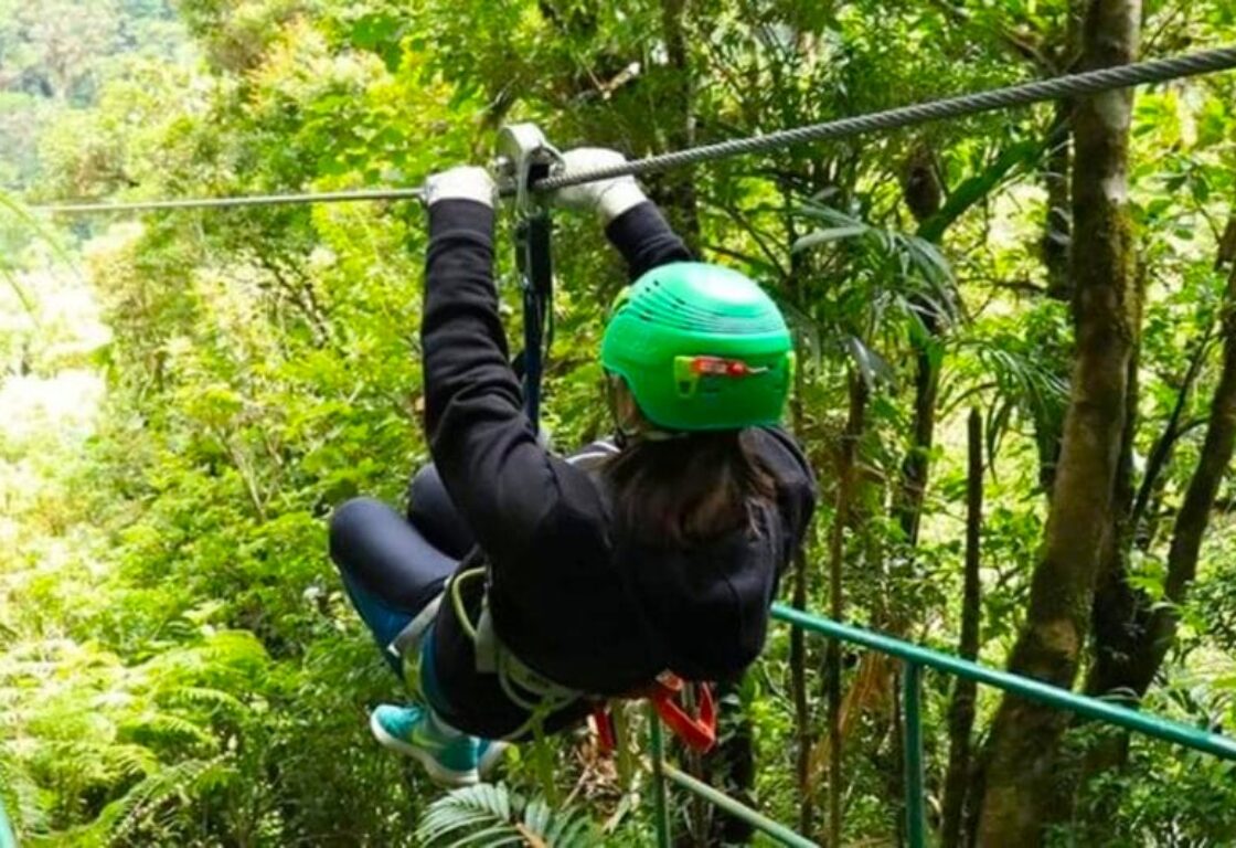 a woman in a black jacket and leggings and green helmet zip lining in Monteverde Cloud Forest