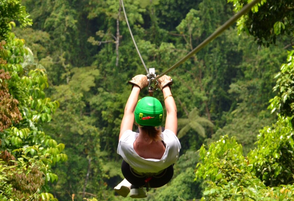 a person in white top and green helmet ziplining into the green forest