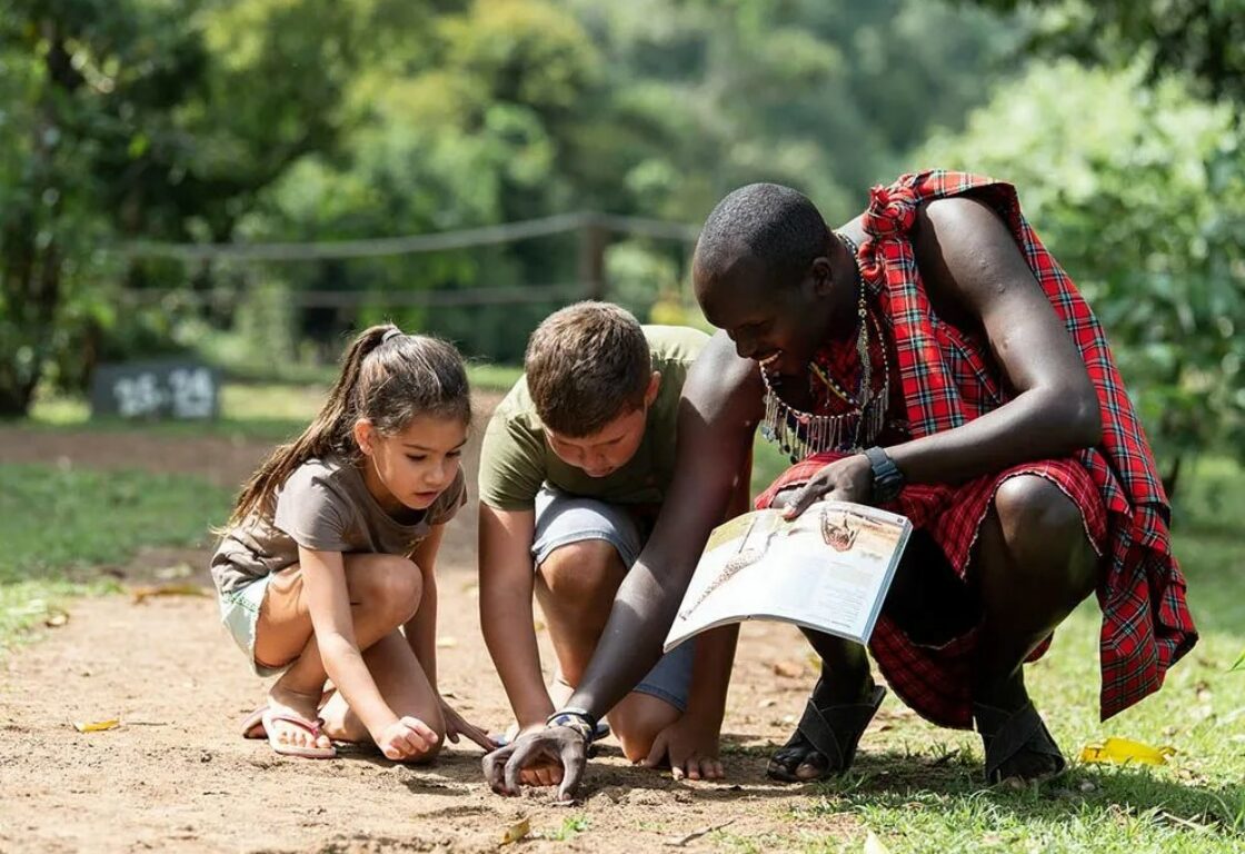 children with a maasai mara man clothed in traditonal fabric with red and blue checks looking at the ground at animals foot prints - children and family friendly safari