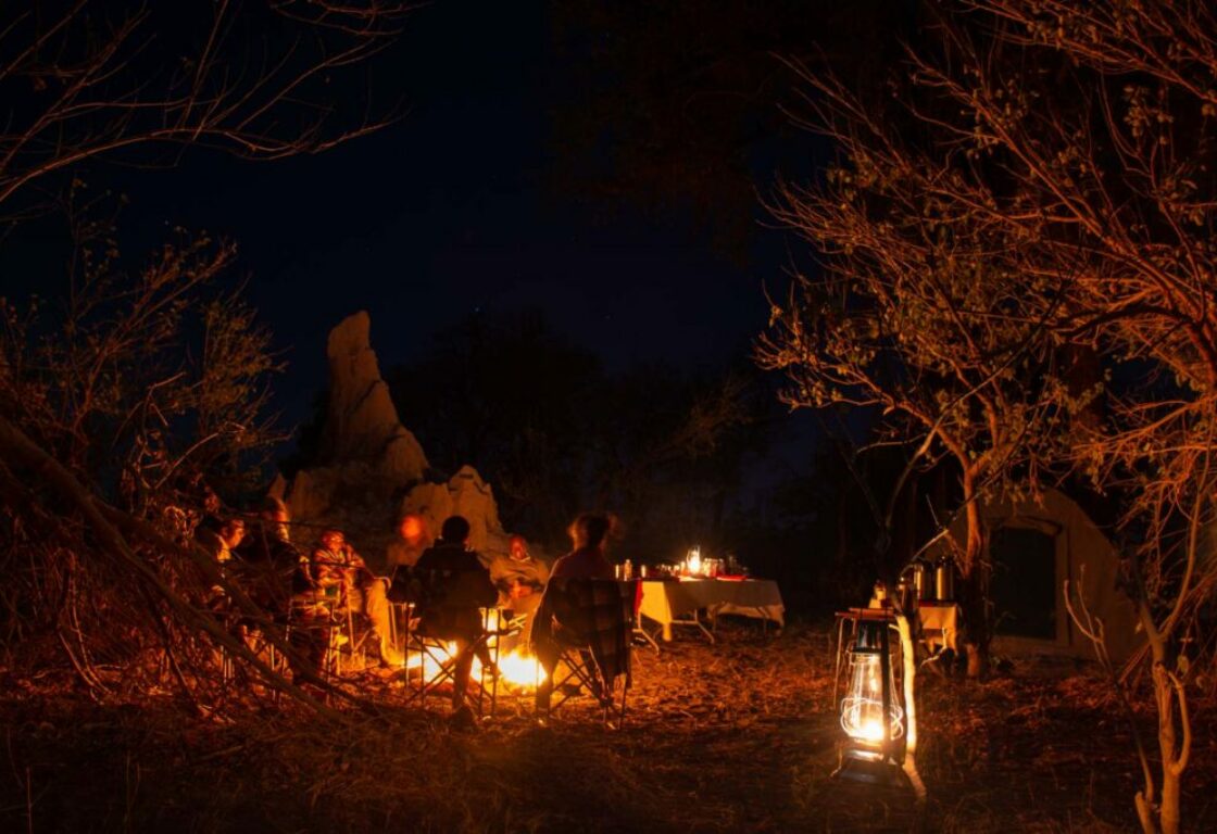 a group of people sitting around a fire in the african bush in the evening