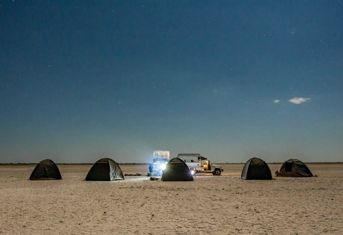 a row of small mobile canvas tents in neutral colours on an extremely bare sandy desert, under a night sky.