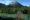A tourist walking toward the camera across a narrow suspension bridge, with turquoise water below and green, forested mountains beyond in Nahuel Huapi National Park, Argentina