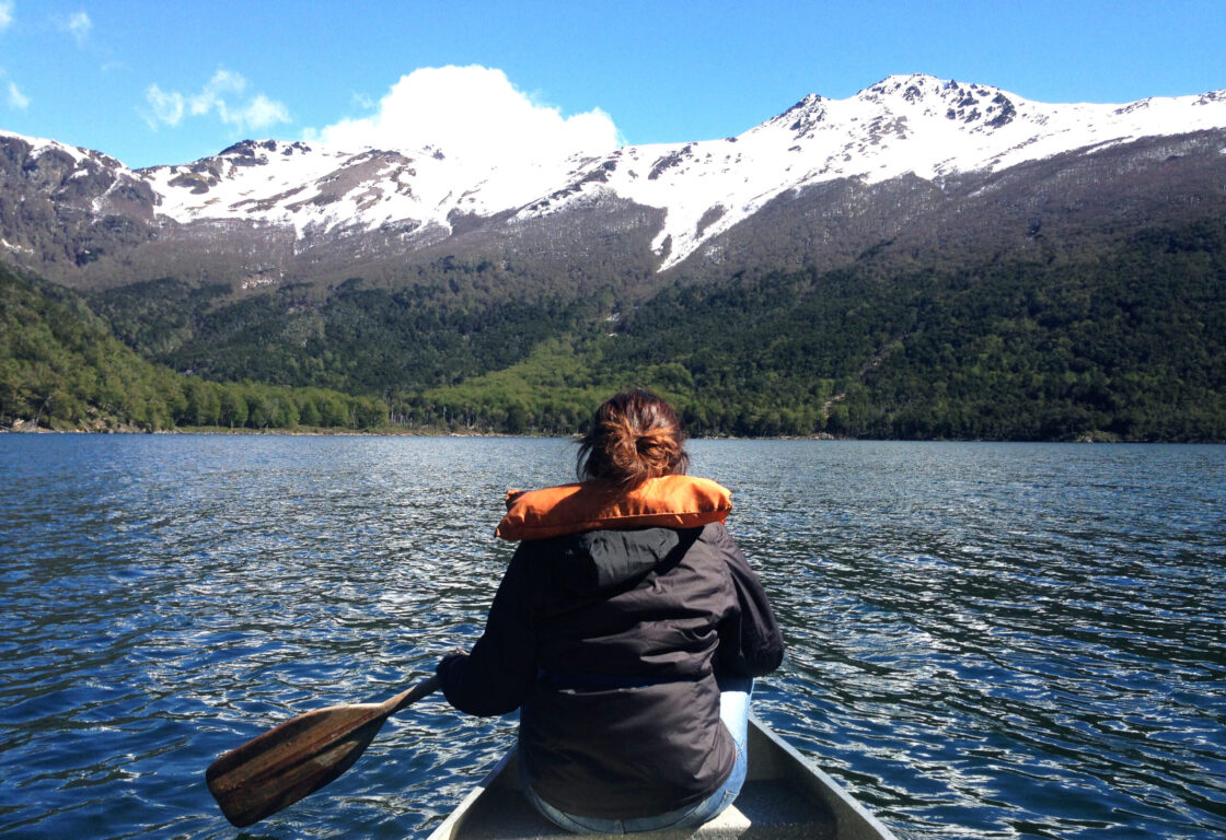 A woman paddling at the front of a canoe on a lake in Argentina's Lake District, with white-capped mountains and forests at the shore