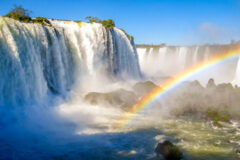 Iguazu Falls pictured from river level, with rainbow in foreground and blue sky behind