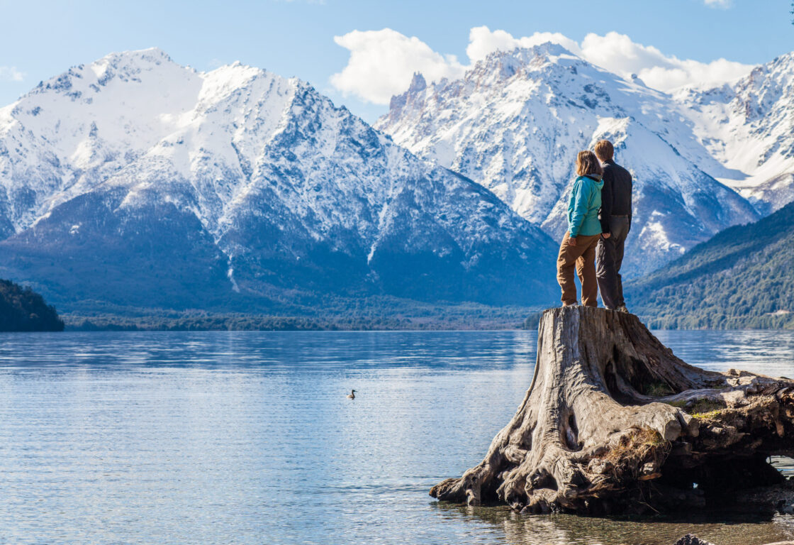 A couple standing on a tree stump jutting over a lake near Bariloche, Argentina, with snowy mountains in the background