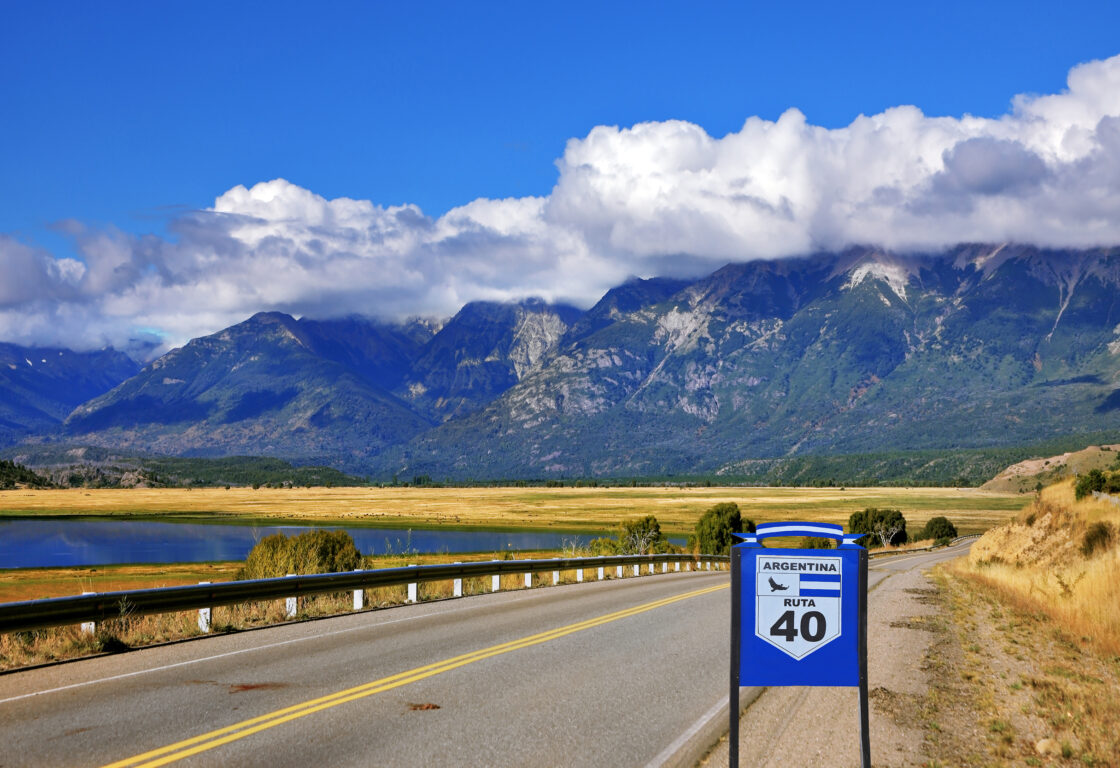 A signposted Ruta 40 stretching out past a lake toward green sloped mountains, Argentinian Patagonia