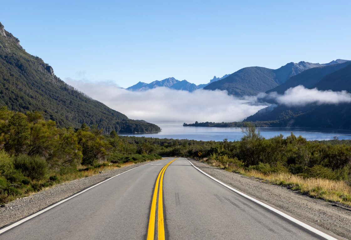 An open road pictured facing toward a calm lake, forests and mountains, with low mist and blue sky, along the Seven Lakes Route, Argentina