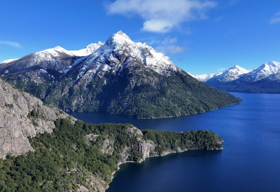 An aerial shot of a blue lake with snow capped grey mountains rising up out of it