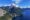 An aerial shot of a blue lake with snow capped grey mountains rising up out of it