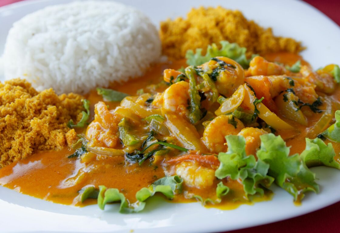 Upclose shot of a white plate with prawns and greens in an orange curry like sauce and a domed shape mound of white rice on the other