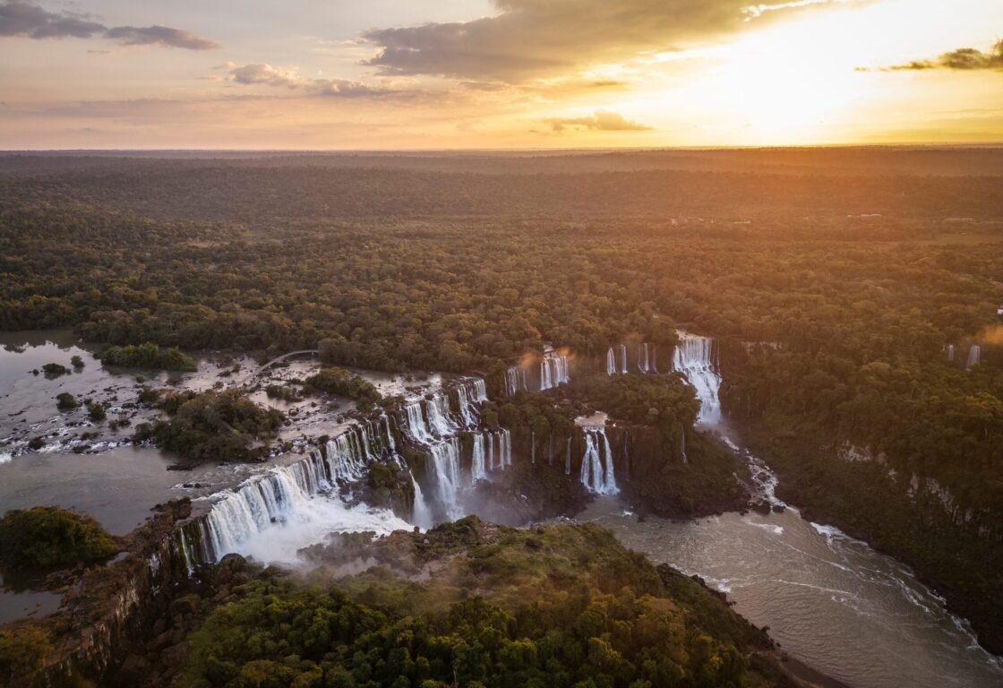 Sunset over Iguazu Falls, a sweeping and dramatic waterfall with miles of lush green rainforest surrounding.