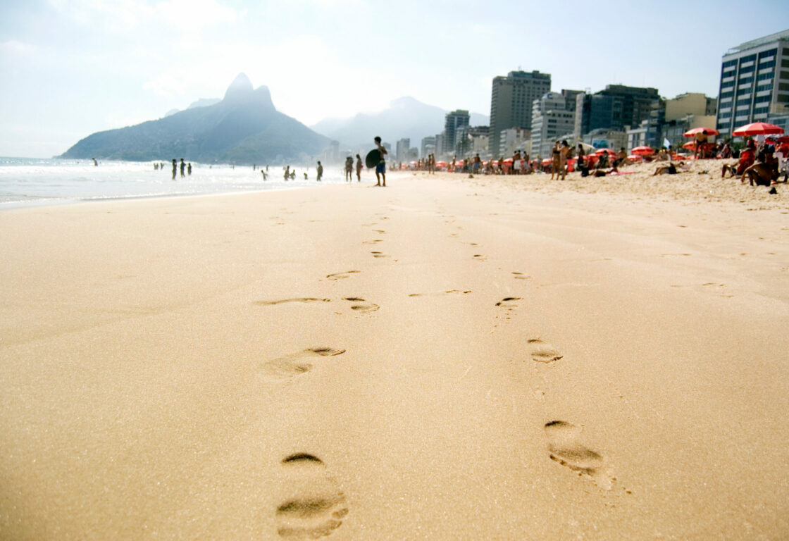 Two sets of footprints along Ipanema Beach in Rio de Janeiro, with beachgoers, cityscape and mountains beyond