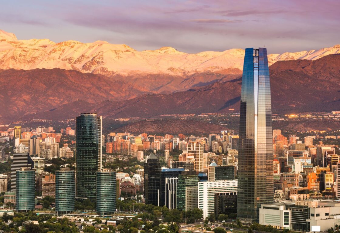 City skyline with a couple ultra modern glass skyscrapers in front and more common high rise buildings lining the city, snow capped mountain range sits at the back of the image, looming over the city