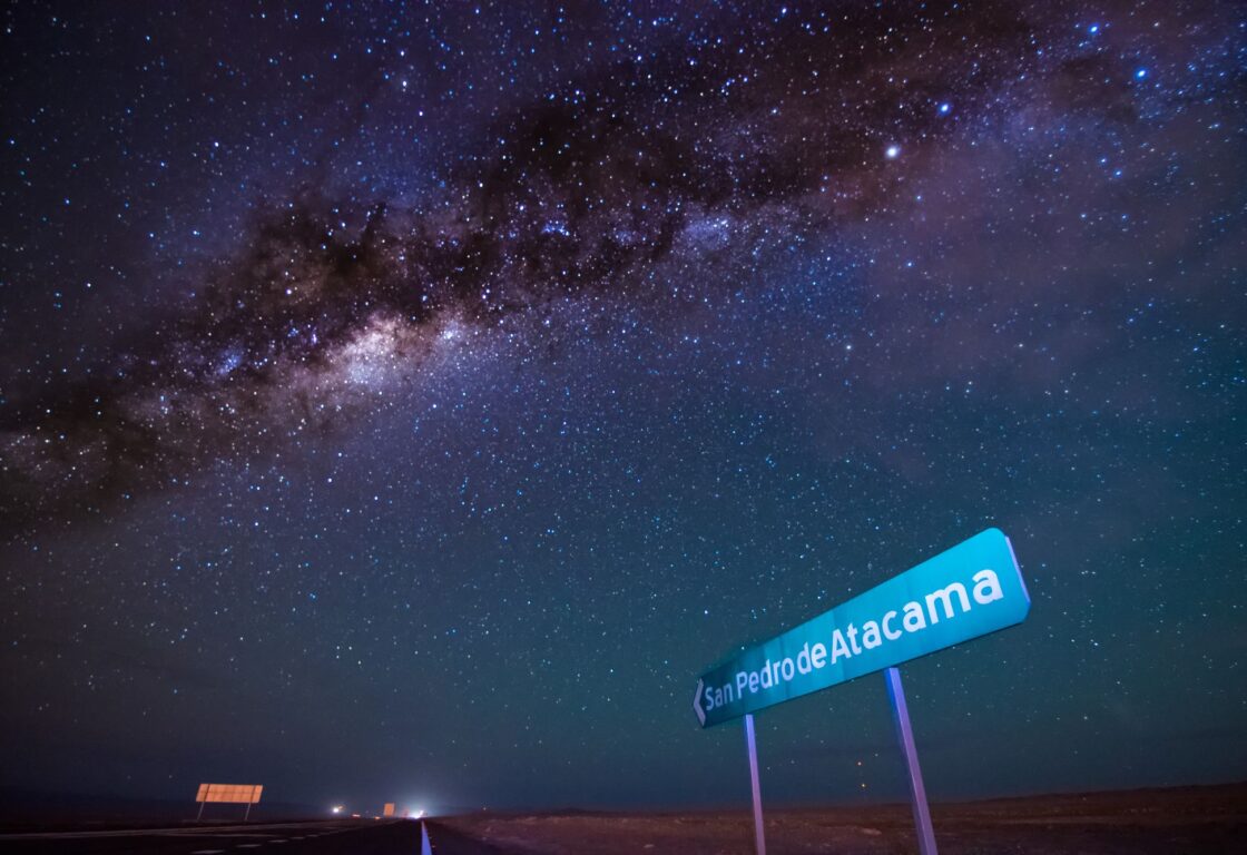 Incredibly vclear ground shot of night sky. Billions of stars dot the sky, with a galaxy. Road sign in bottom right, sgnalling the way to San Pero De Atacama