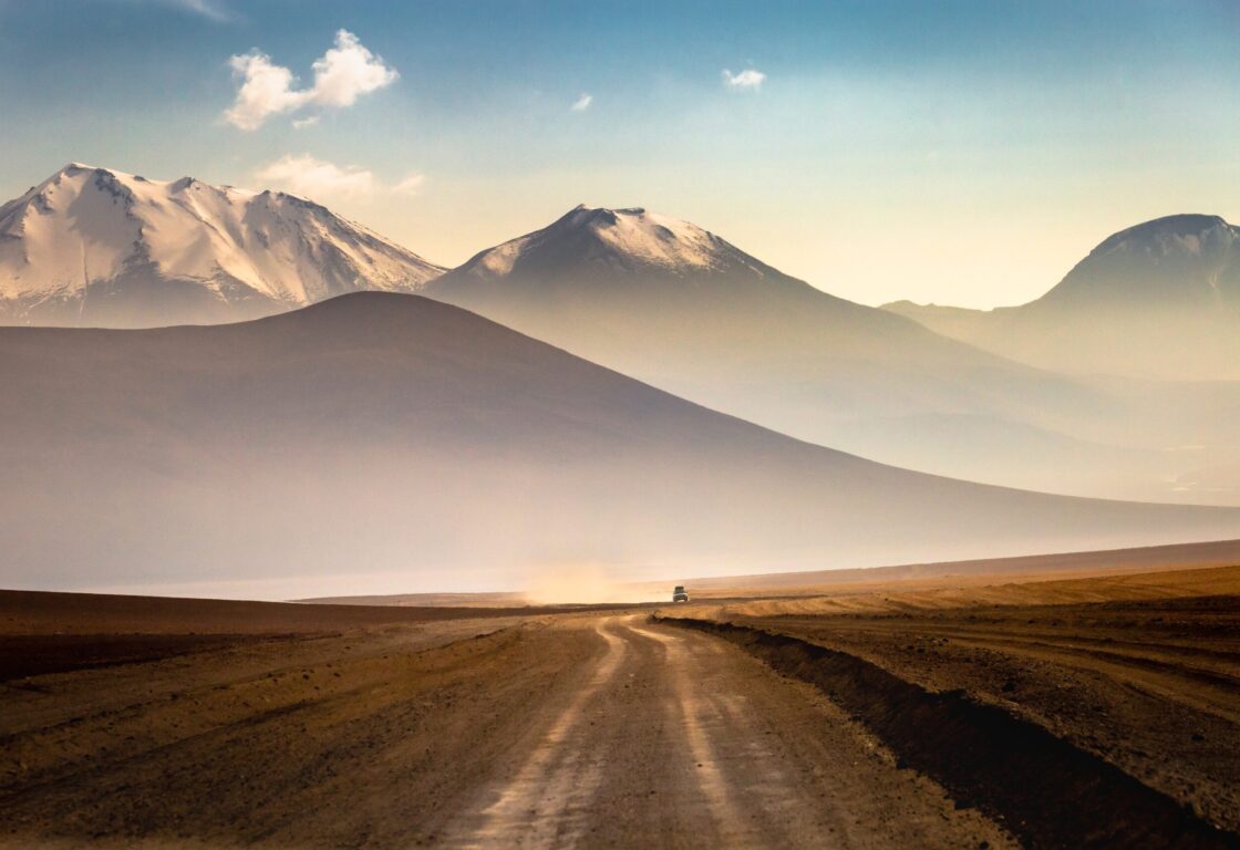 A vast plain is divided by car tracks, forming a road, whilst dramatic snow capped mountains, rise up in the distance