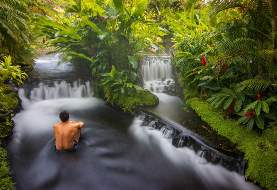 A shirtless man sits in a serene stream, surrounded by lush greenery and gentle waterfalls, creating a tranquil natural setting.