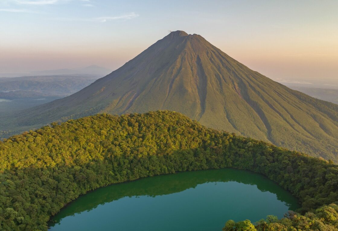 A blue lake is surrounded by lush green rainforest hills, whilst in the distance a triangular volcano rises up