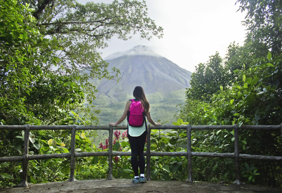 Female hiker stands at a lookout point, amidst tropical jungle trees, to an opening where the Arenal Volcano, a symmetrical stratovolcano rises up